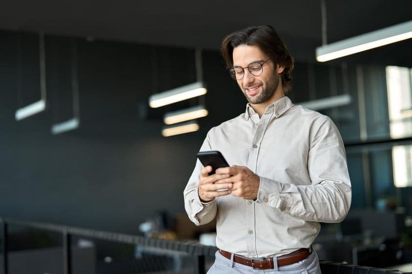 Fiatal európai üzletember mobiltelefont használ munkahelyén, hivatalban állva.