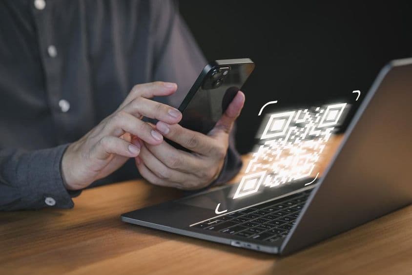 Egy személy okos telefonnal olvassa be a laptop képernyőjén megjelenő QR-kódot.